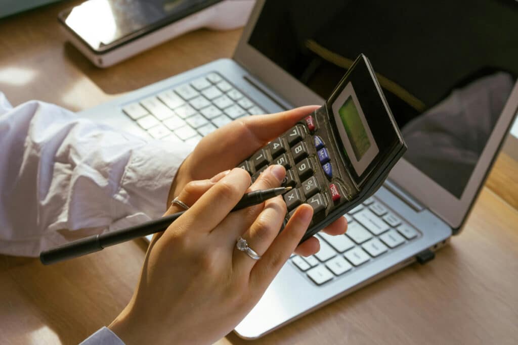 Hands using a calculator at a desk with laptop and notes, showing contractor estimating software workflow.